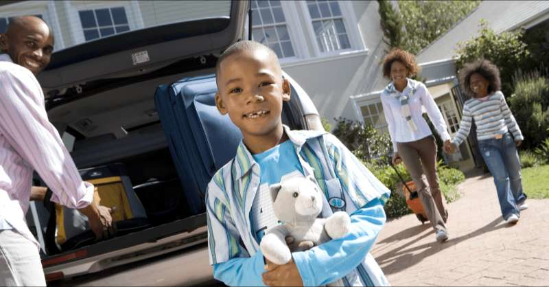 A family packs their car and prepares to leave on a trip as a little boy holding a Teddy bear looks into the camera