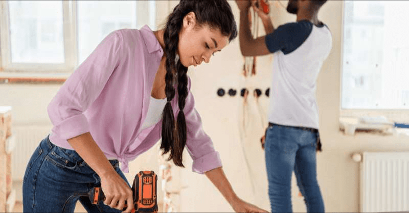 A woman works a jigsaw as her partner is seen working in the background as they work on their home renovations