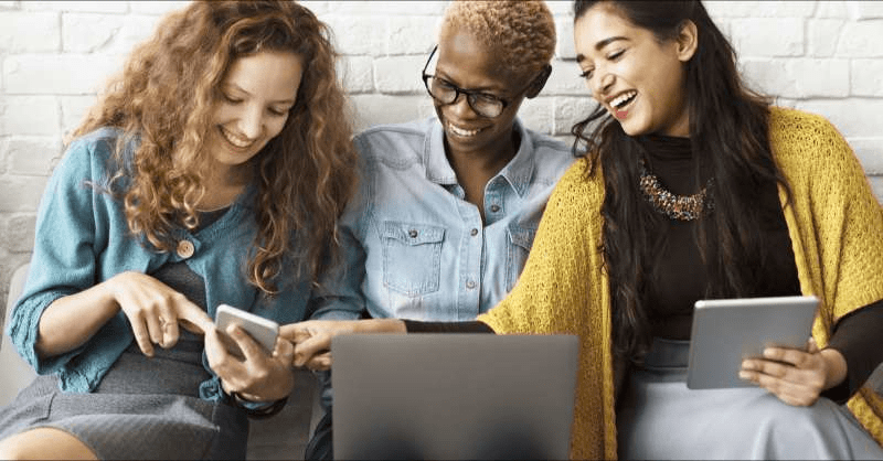 A small group of women laugh and smile and they look at their electronic devices