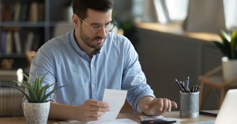 A young man sits at his desk with his laptop and calculator, and is working on revising his budget for the new year.