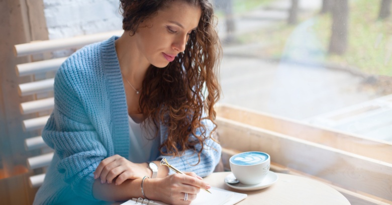 A young woman has a warm cup of coffee next to her on the table and is ready to write in her notebook a few ways to save money.