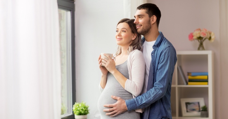 A young man is hugging his pregnant wife who is holding a cup of coffee. They are both looking out the window and enjoying the moment.