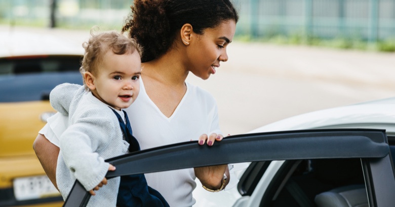 A mother holds a toddler in her arms as she prepares to put the child in her car seat in the back seat of the car.