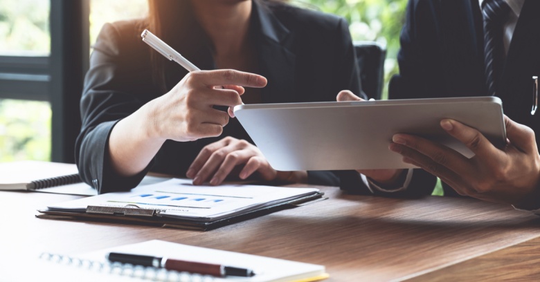 Two people are shown from the shoulders down. They are looking at investment charts and a computer to decide how much of a tax refund to put towards investments