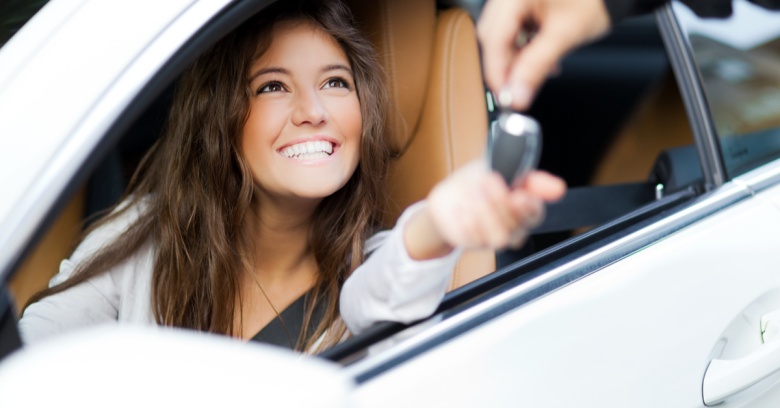 A smiling woman sitting in the driver's seat of a car smiles as keys to her new car are handed to her. She is happy to be building her credit with her auto loan.