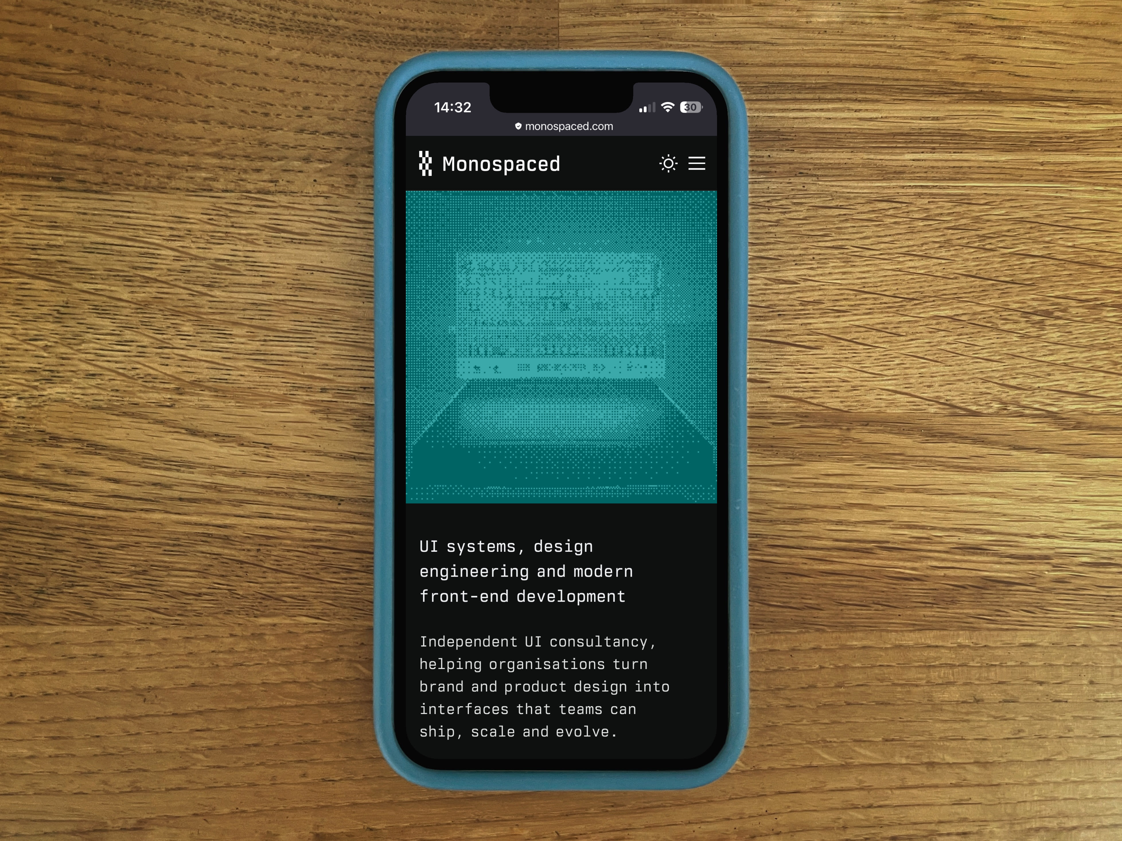 A phone on an oak table showing the monospaced.com homepage: a cyan halftone image above the strapline and introduction in monospaced type.