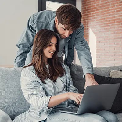A couple using a laptop together in their living room. One person is pointing at something on the laptop.