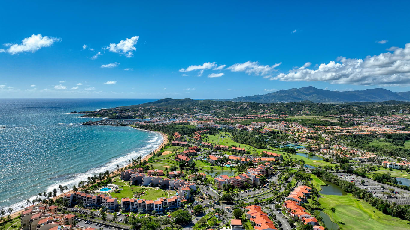 Aerial view of Palmas del Mar, Puerto Rico: a coastal resort lush greenery, golf courses, and the coastline meeting the blue ocean, surrounded by mountains under a bright blue sky.