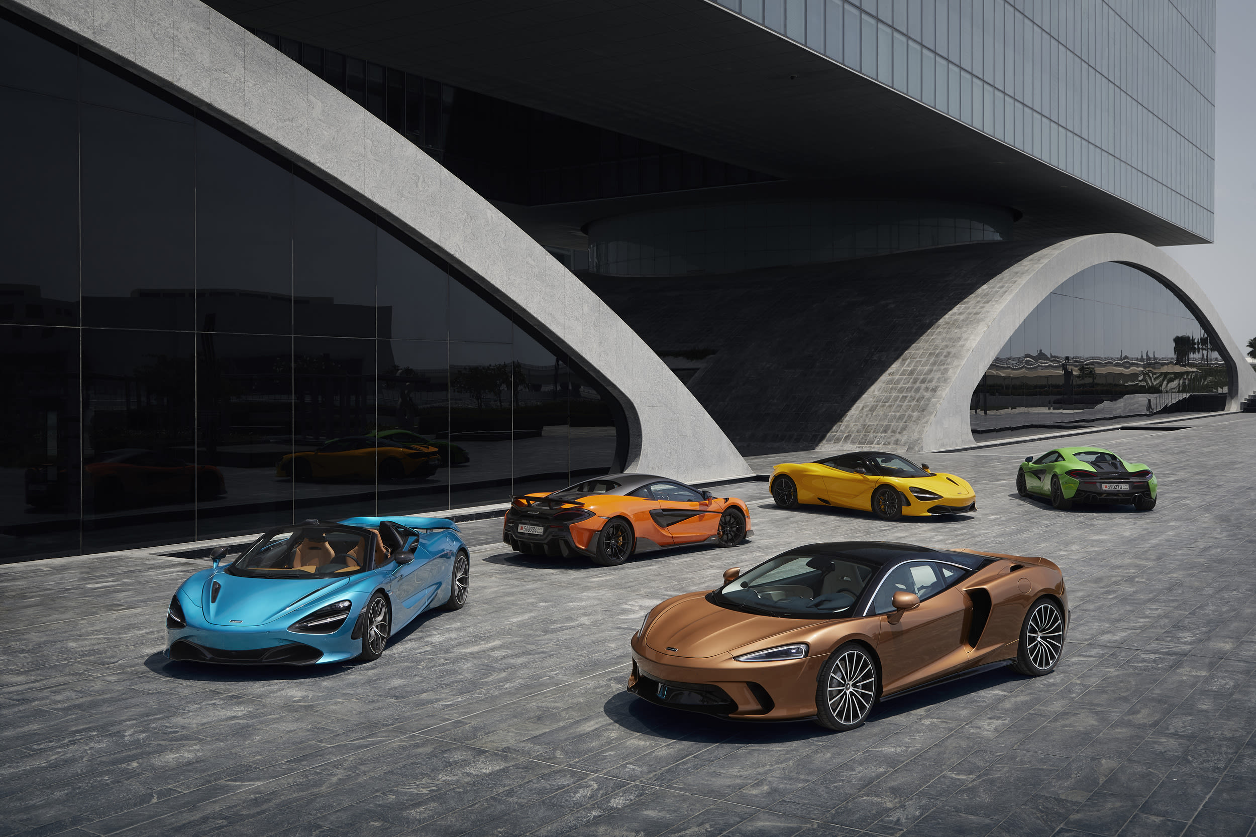 Five McLaren sports cars parked outside a McLaren retailer, with a bronze GT in the foreground