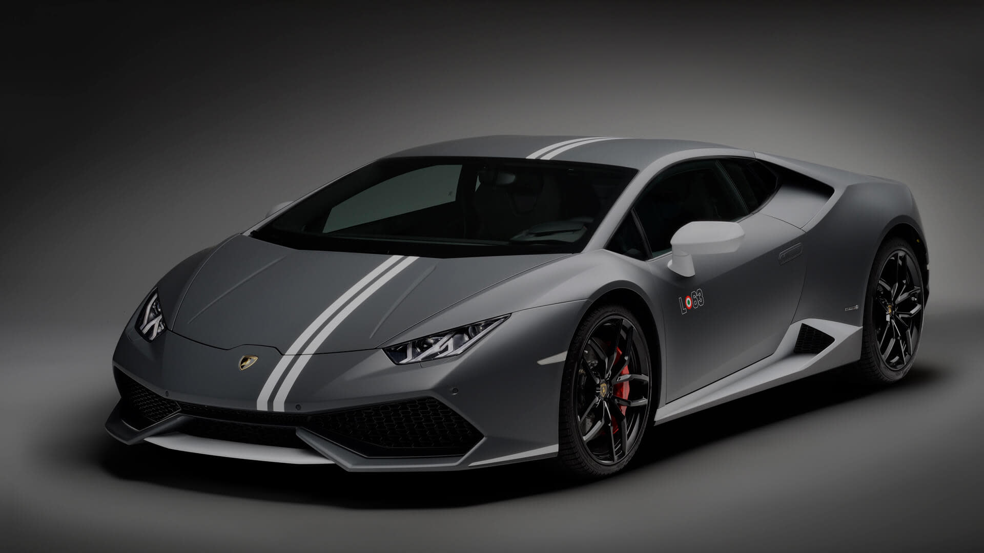 Vista frontale di tre quarti di una Lamborghini Huracán Avio in grigio opaco con doppie strisce, parcheggiata su un asfalto sotto un cielo limpido