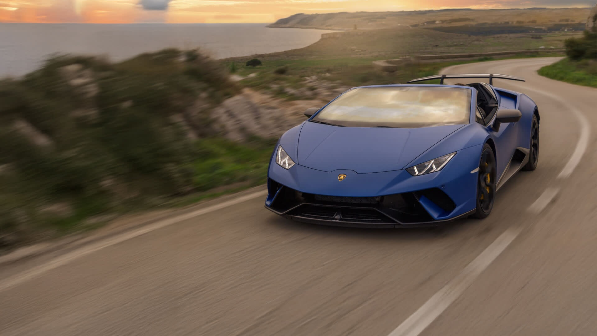 Dreiviertelansicht eines Lamborghini Huracán Performante Spyder, der mit offenem Verdeck eine Küstenstraße entlangfährt und dabei Geschwindigkeit und Eleganz betont.
