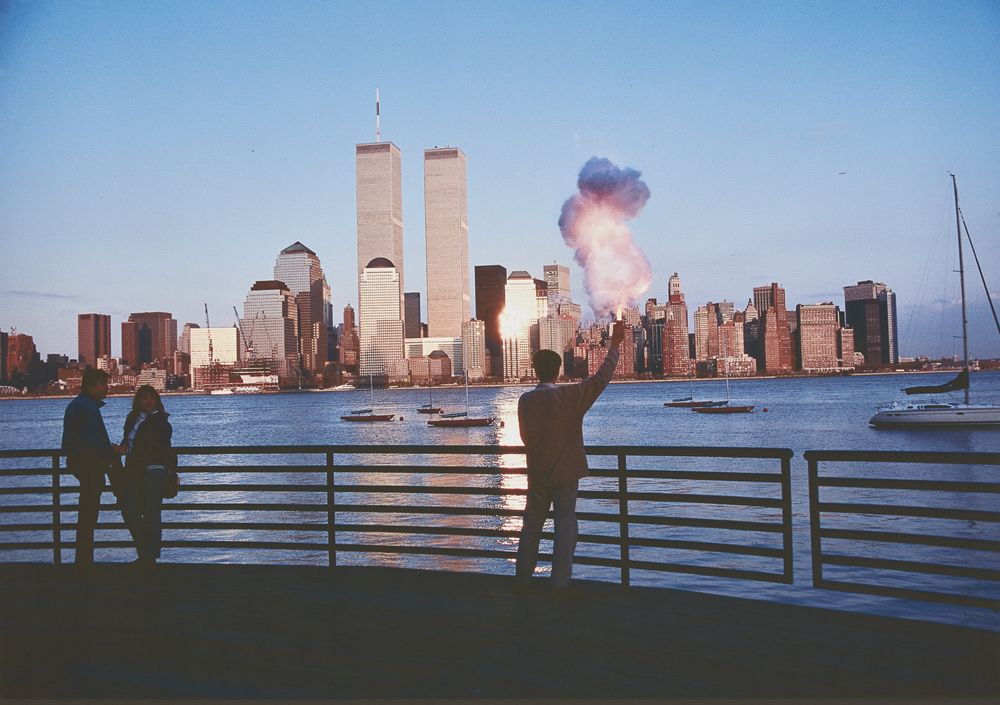 Photograph of a man standing on a promenade with his back turned, against the Manhattan skyline across the water. Puffs of smoke rise from his raised right hand. Two figures on the viewer's left turn their heads towards the man.