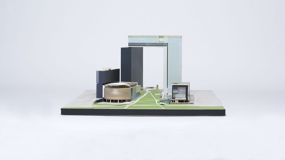 Cardboard and acrylic architectural model of a building complex. In the centre, a rectangular grey building on our left and an inverted L-shaped pale blue building form an open door that towers over the complex. Buildings stand on both sides of a green corridor running beneath the open door.