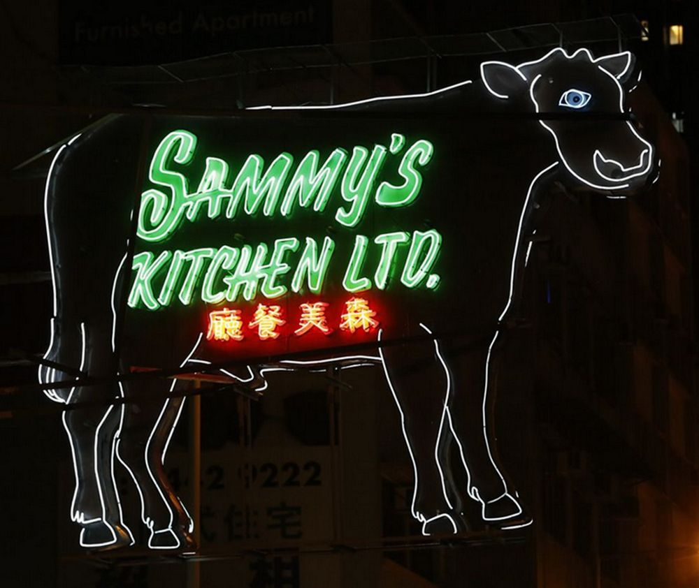 A neon sign in the shape of a cow with a white outline, viewed against a dark night sky. The words ‘Sammy’s Kitchen Ltd.’ in green English and red Chinese lettering are displayed inside the cow.