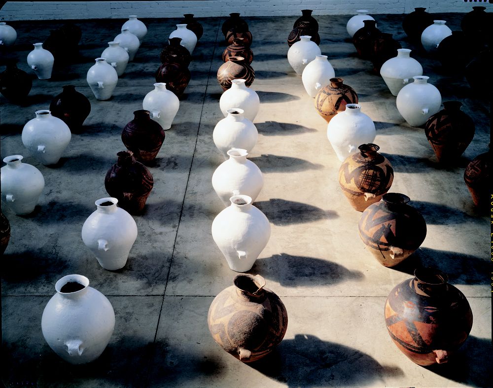 Installation consisting of dozens of individual clay pots that are narrow at the open top, bulbous in the middle, tapered at the base, and are arranged in rows. Some are terracotta orange adorned in black, while others have been painted white.