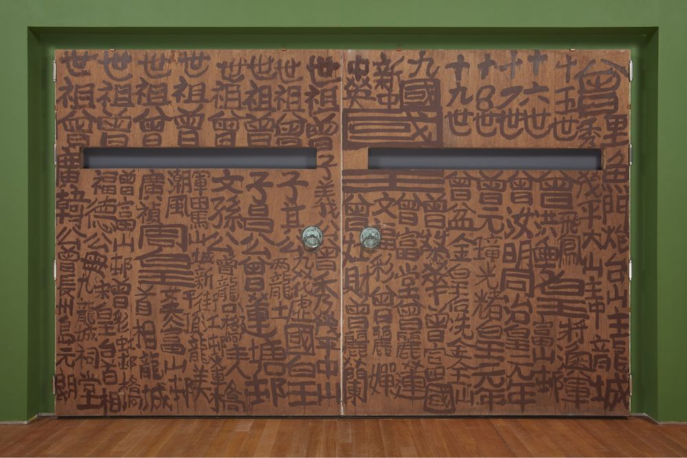 墨水木門裝置作品豎立於一面凹入的綠色牆壁前,門上設有兩個門環,上半部分有兩條細長的橫向長方形穿孔。兩扇門的表面以黑色墨水寫滿大小不一的方塊狀中文字,並重複展示幾個中文字,包括「國」和「皇」。