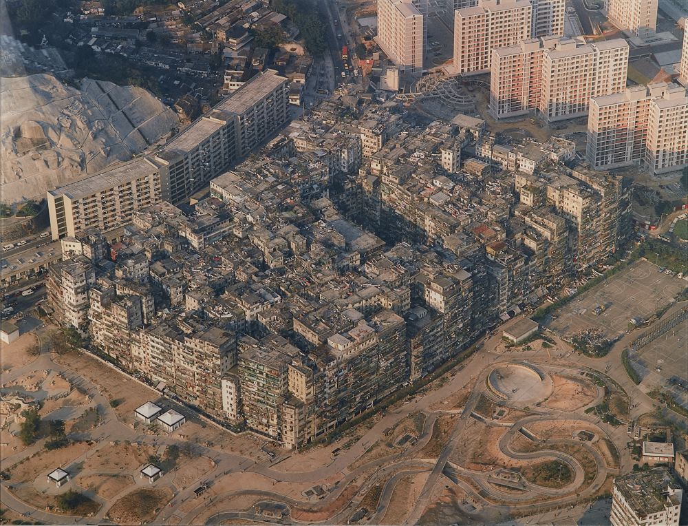 Photograph of a rectangular mass of very tightly packed tall buildings viewed from above. The buildings are surrounded by empty land covered in roads and a group of more sparsely spread out high-rises.