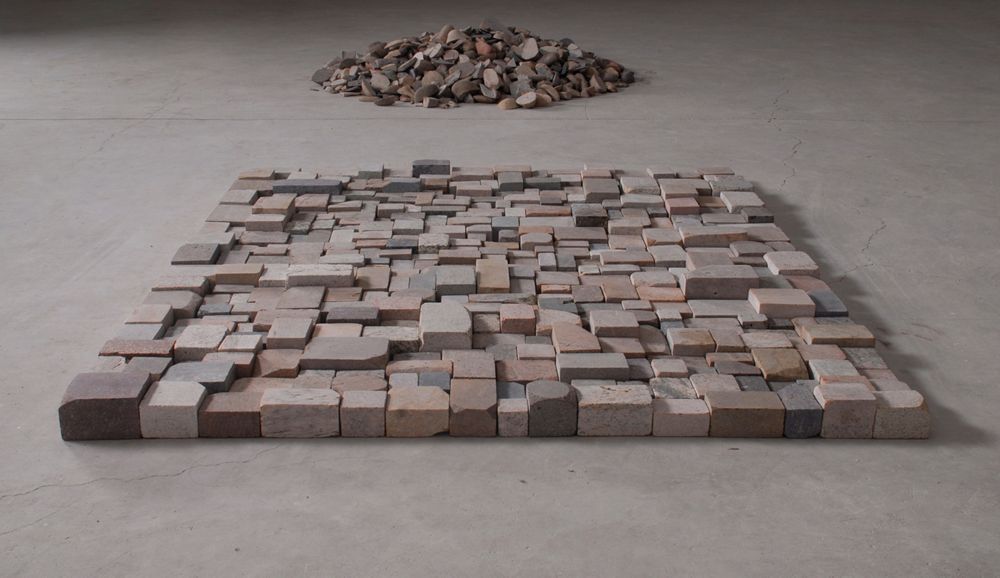 The installation consists of two arrangements of pink, grey, and rust cobblestones on the floor. One of the arrangements, which lie further in the distance, has irregular stones piled to form a circle, while the one in the foreground is a neat square assembled by cuboids in different sizes.