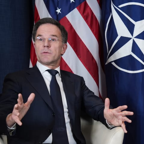 Mark Rutte (left) and Donald Trump sit next to each other in front of flags for the U.S. and NATO. Trump looks at Rutte as he speaks.