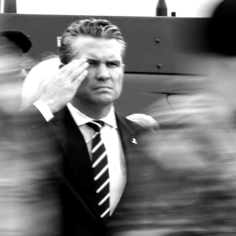 Pete Hegseth (far left) and Dan Caine (far right) salute as people in military uniforms move in front of them, blurred in motion. The photo is in black and white.