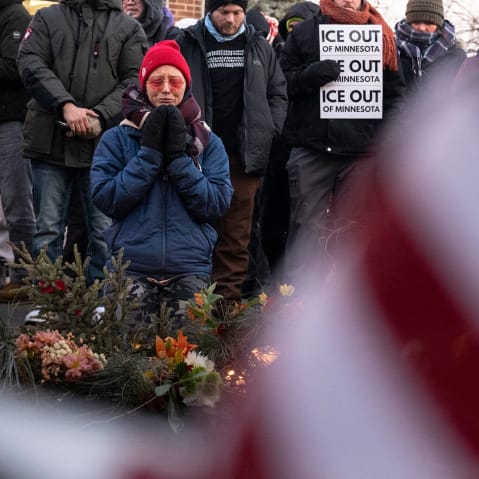 Part of an American flag fills the frame, blurred, in the foreground. On the left of the frame, a person kneels by a memorial of flowers.