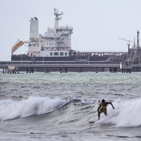 A man is seen from behind as he surfs in front of an oil tanker in the distance.