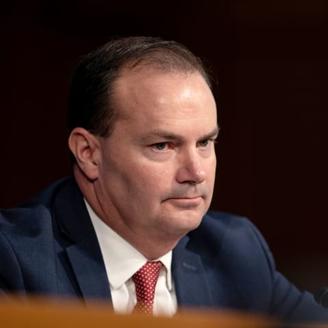 Mike Lee looks on during a hearing. The background is completely black.