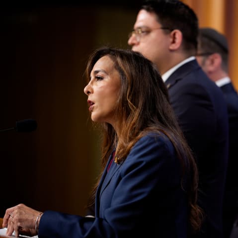 Harmeet Dhillon speaks at a podium. This is a profile shot. She is surrounded by aides.