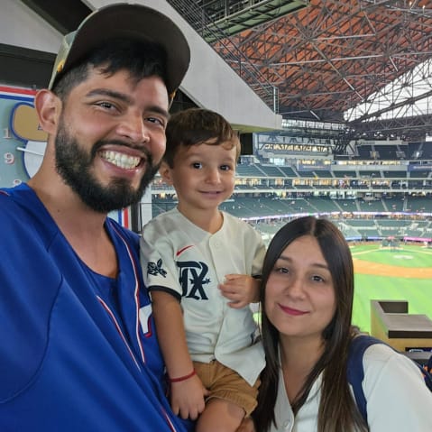 A man, woman, and small child, all in baseball jerseys. Behind them is a baseball field.
