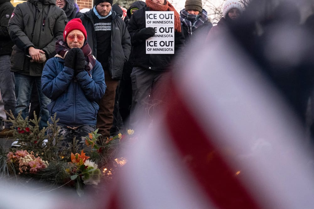 Part of an American flag fills the frame, blurred, in the foreground. On the left of the frame, a person kneels by a memorial of flowers.