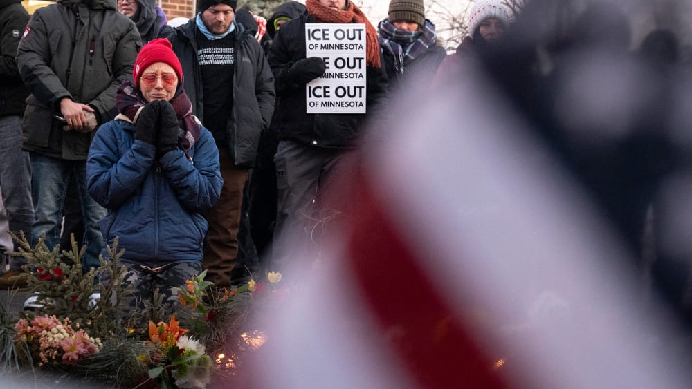 Part of an American flag fills the frame, blurred, in the foreground. On the left of the frame, a person kneels by a memorial of flowers.