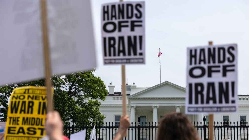 Out of focus in the foreground, people hold picket signs that read "Hands Off Iran!" In focus in the background is the White House.