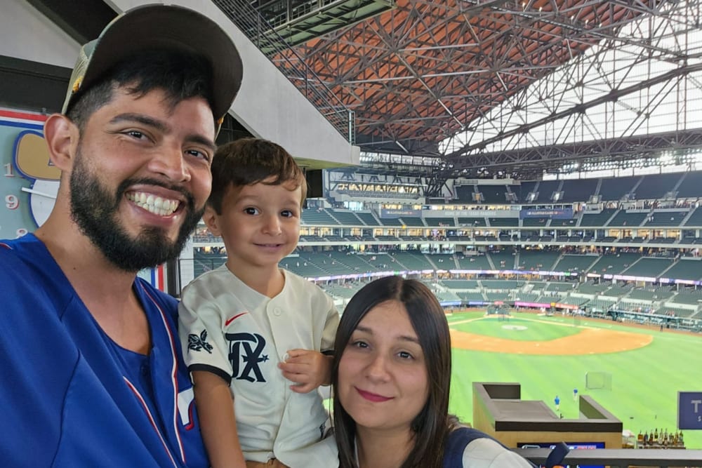 A man, woman, and small child, all in baseball jerseys. Behind them is a baseball field.