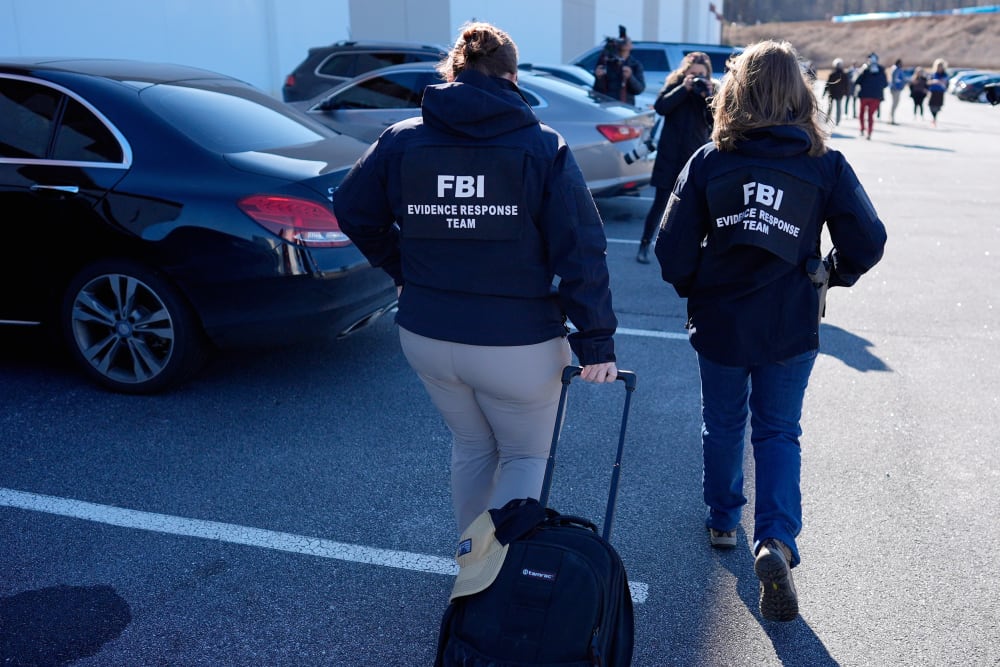Two FBI agents walk in a parking lot, one dragging a bag.