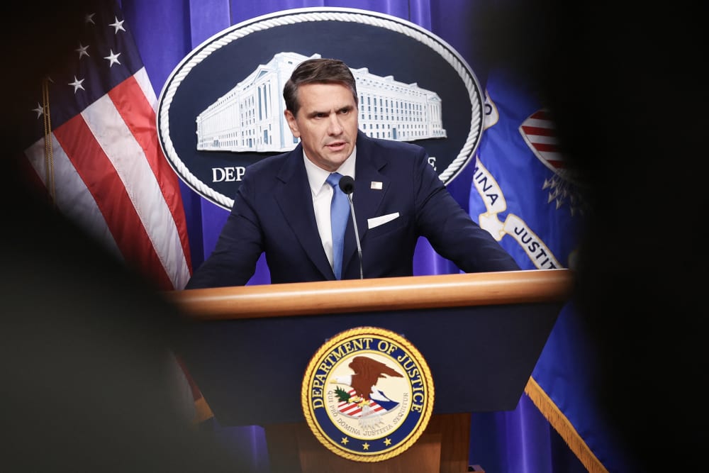 Todd Blanche is seen speaking at the podium through other people in the room.