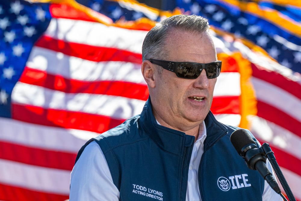 Todd Lyons speaks at a podium outside during daytime.