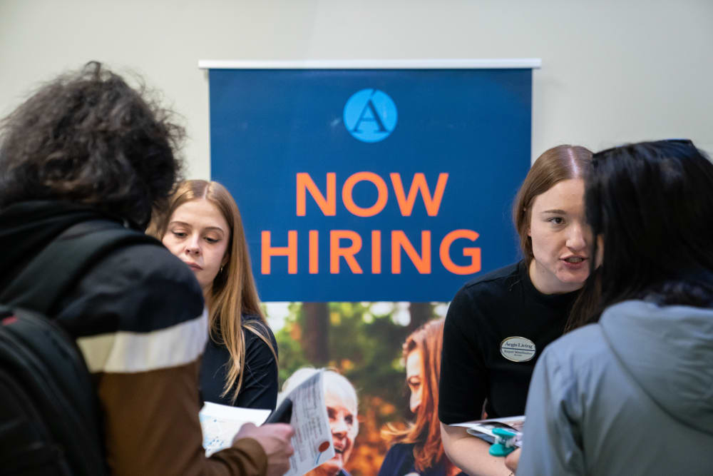 Two people talking with two job seekers in front of a &ldquo;Now Hiring&rdquo; sign.