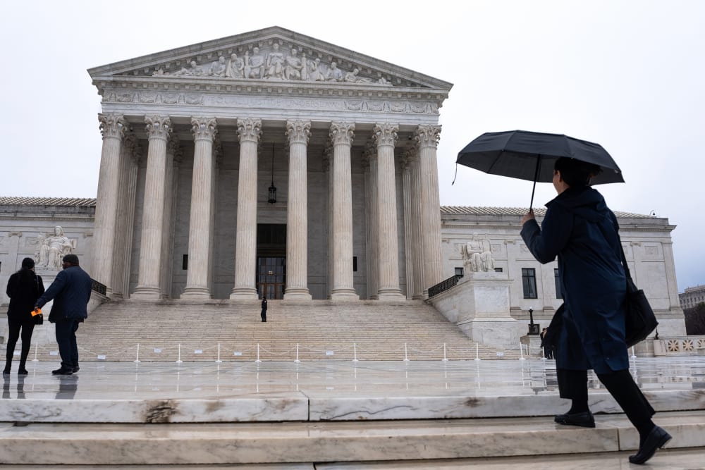 A person with a black umbrella walks past the SCOTUS building in the rain.