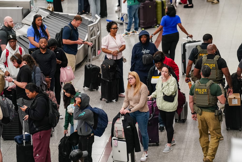 People wait in line at the baggage claim area as three ICE agents walk around.