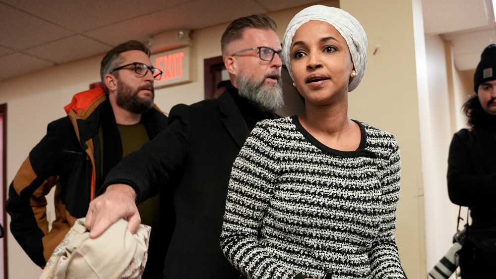 Ilhan Omar walks into a room for a town hall meeting, followed by two men who seem to be security personnel.