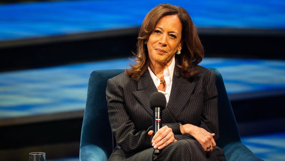 Kamala Harris sits at the stage while smiling and her hands are crossed over her legs.