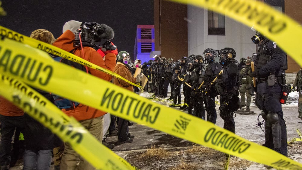 People and law enforcement stand behind strands of yellow caution tape.