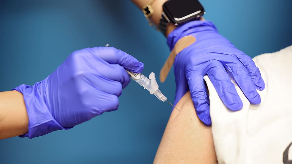 The hands of a person in medical gloves gives a shot to a person whose shoulder is visible.