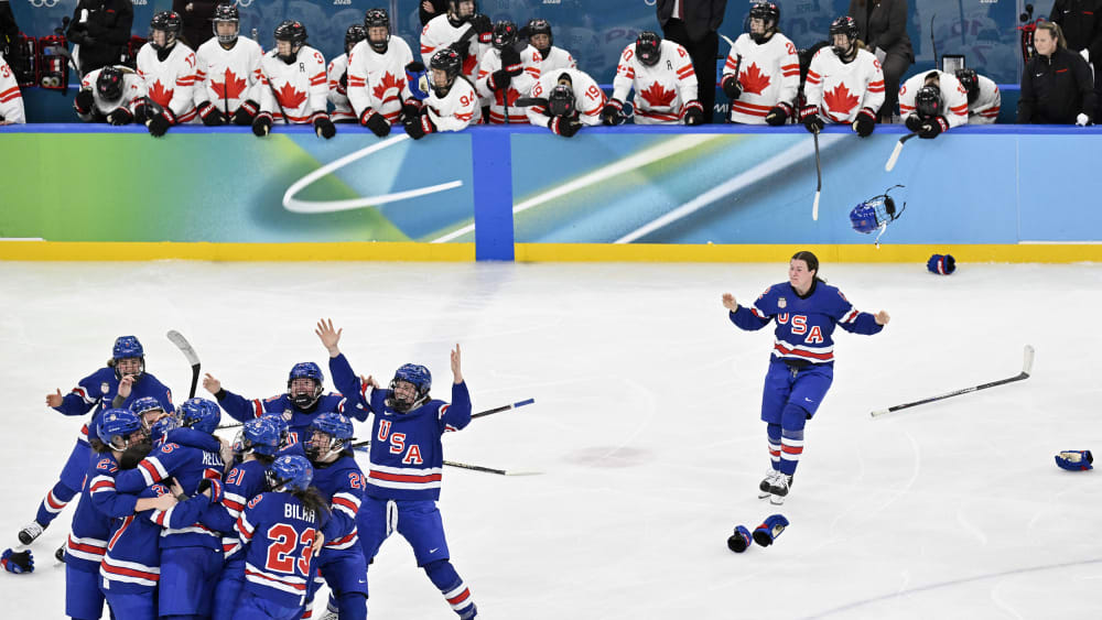 A group of US hockey players are huddled on the left side of the frame as one player runs toward them in celebration.