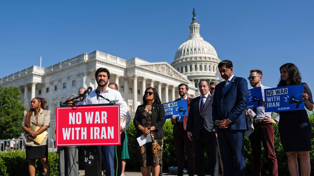 Greg Casar stands behind a podium that has a red sign that says "NO WAR WITH IRAN." He is surrounded by other people, and in the background is the Capitol.
