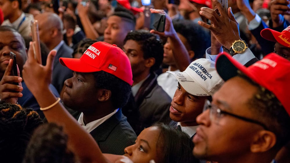 Audience members, most of them Black, wear MAGA hats and hold up their phones to take photos.