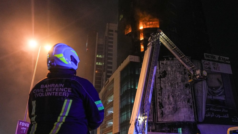 A person in a hard helmet with a jacket that says "Bahrain Civil Defence (Volunteer)" looks up at a building which is on fire.
