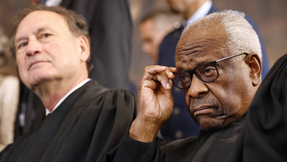 Clarence Thomas adjusts his glasses as Samuel Alito leans back in the background.