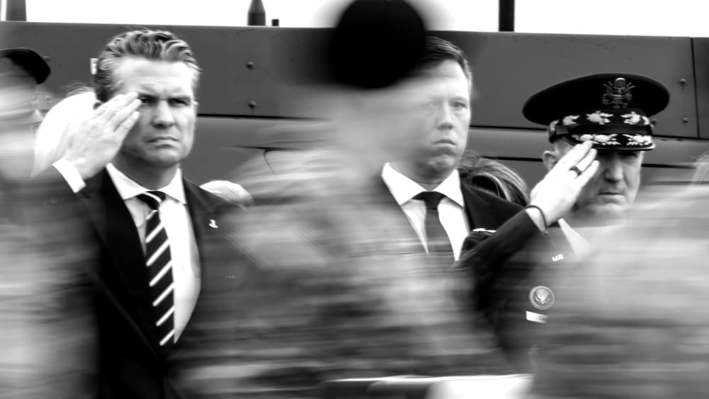 Pete Hegseth (far left) and Dan Caine (far right) salute as people in military uniforms move in front of them, blurred in motion. The photo is in black and white.