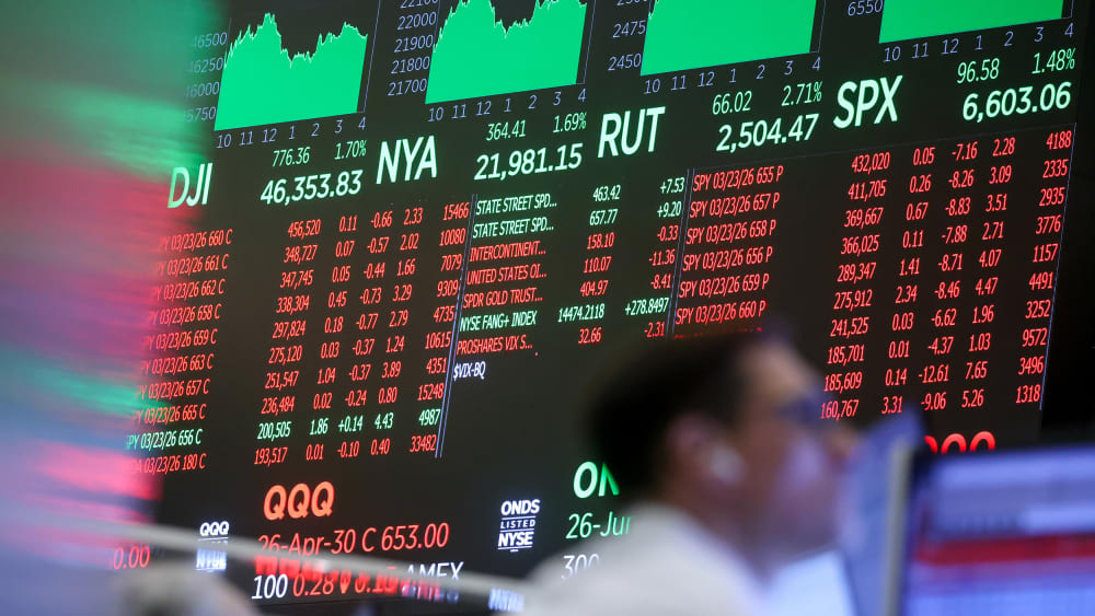 Stock exchange monitors can be seen in the background in focus as two men work in the foreground.
